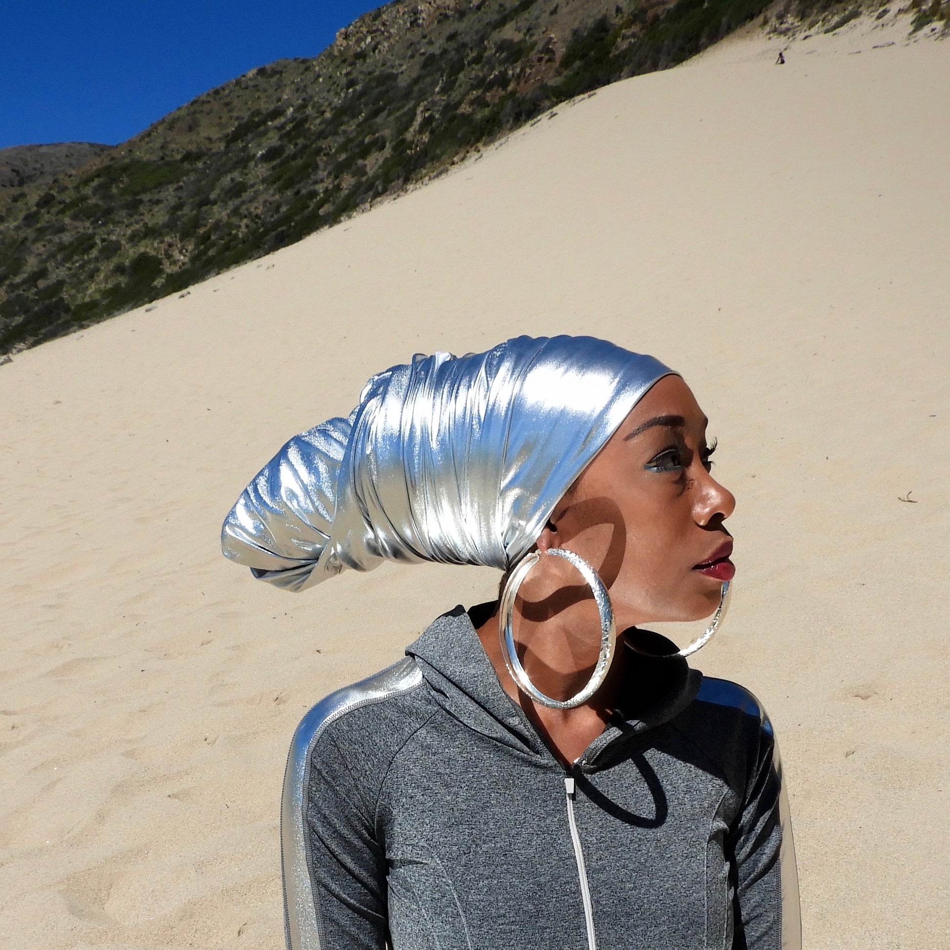 model wearing stretchy metallic silver head wrap in the sand. head wrap style in low cone style. model is sitting in sand with mountains in background